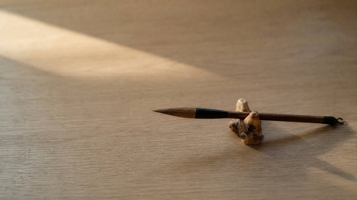 A calligraphy brush resting across a natural bamboo root brush rest on a pale wooden desk in soft window light