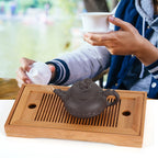 Hands lifting a gaiwan above a bamboo gongfu tea tray outdoors