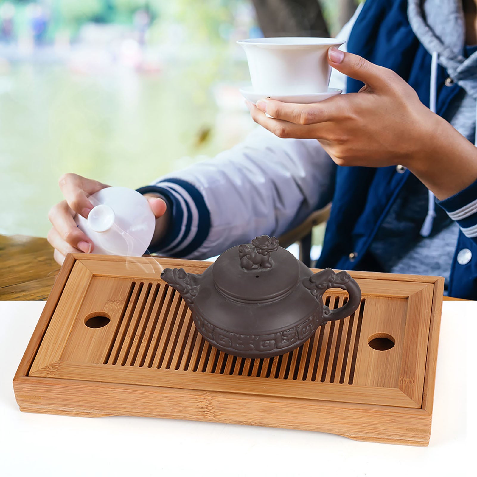 Hands lifting a gaiwan above a bamboo gongfu tea tray outdoors