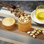 Small bamboo canister holding pistachios next to its round bamboo lid and a cup of tea, Serein Home