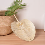 Woven palm leaf hand fan leaning against a seagrass basket on a wooden floor